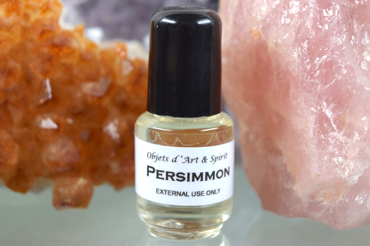A bottle of Persimmon Oil with a label that reads 'Persimmon'. The bottle is placed in front of a crystal with a similar color to the oil.